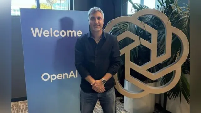 אדם עומד ליד שלט "Welcome" של OpenAI ולצידו הלוגו המעוגל של החברה, במשרד
