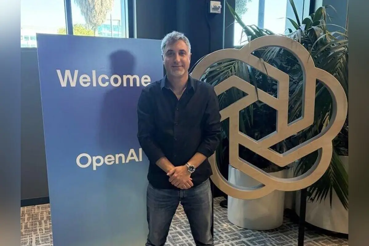 אדם עומד ליד שלט "Welcome" של OpenAI ולצידו הלוגו המעוגל של החברה, במשרד